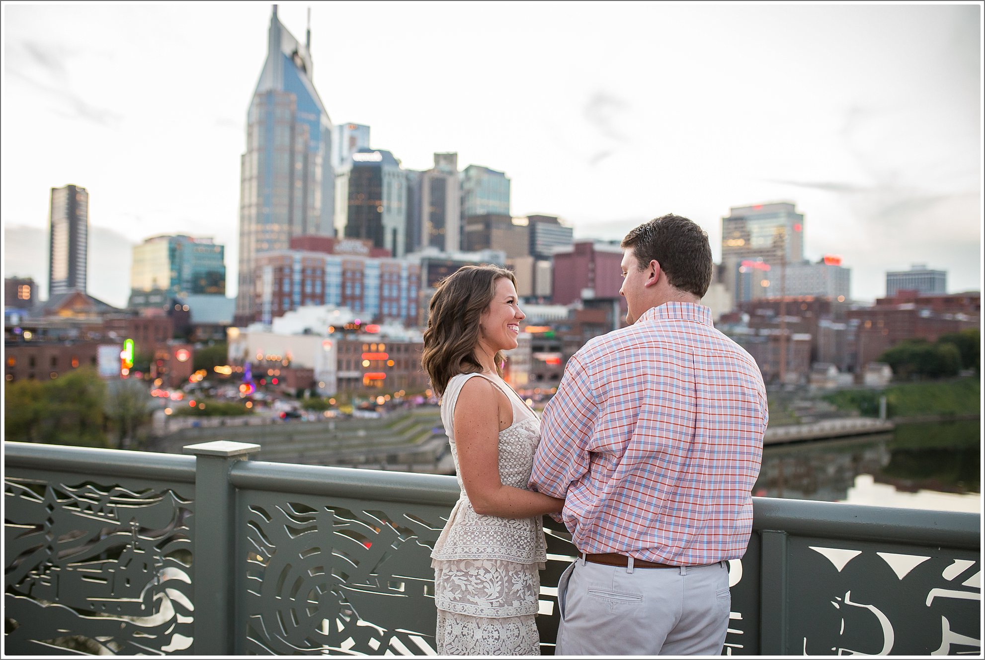 A beautiful engaged couple celebrate their love with an engagement session in Nashville on the pedestrian bridge at sunset with the city skyline lit up in the background.