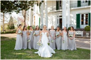 blonde bride in white gown and bridesmaids in grey dresses in front of Riverwood Mansion Nashville