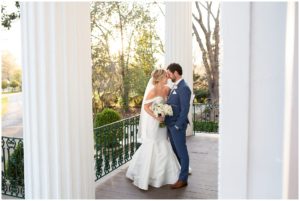 bride in off the shoulder gown and groom in navy tux smiling around tall white columns