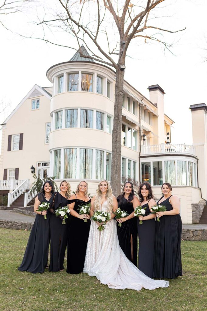 Bridesmaids in elegant black gowns and a bride in a glittery bridal gown and long blonde hair have their portrait taken together with mansion at the Estate at Cherokee Dock as the background. 