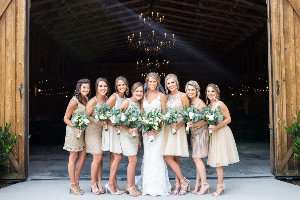 Beautiful bridesmaids in neutral short length bridesmaids gowns accompany a gorgeous bride in a lace gown as they pose for a portrait for Melanie Dunn Photography at Saddle Woods Farm.