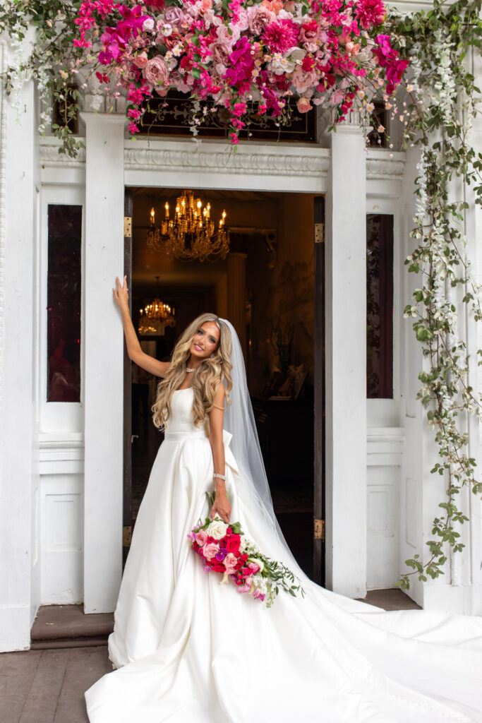 gorgeous tall thin blonde bride in the front doorway at Riverwood Mansion with her bright pink floral display
